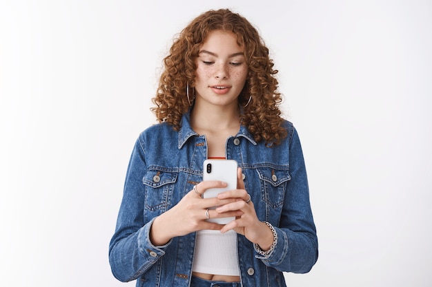 Resta fermo un altro colpo. focalizzato carino hipster rossa ragazza lentiggini che tiene lo smartphone guarda attentamente il display del telefono scattando foto amico catturando immagine per blog internet, in piedi sfondo bianco