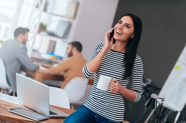 Rimanere connesso. bella giovane donna sorridente che tiene una tazza di caffè e parla al telefono cellulare mentre i suoi colleghi lavorano in background