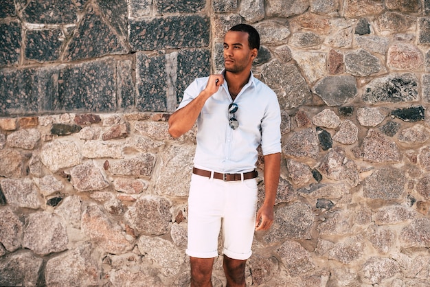 Staying cool and stylish. Handsome young African man in smart casual clothes adjusting his shirt collar while standing against the stoned wall outdoors