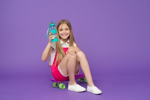 Staying hydrated Girl happy face holds with water bottle while sits skateboard violet background Kid girl keeps staying hydrated Body hydration concept Girl cares about health and water balance