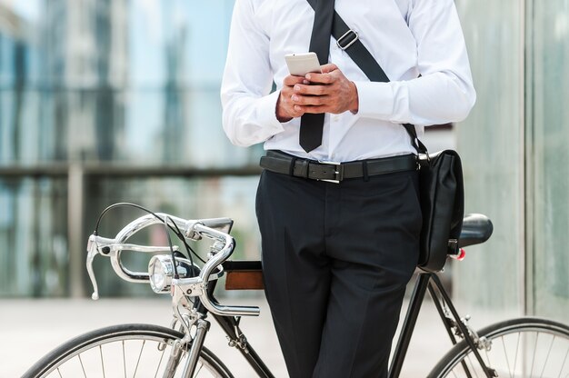 Staying in touch. Close-up of young businessman holding mobile phone while leaning at his bicycle with office building in the background