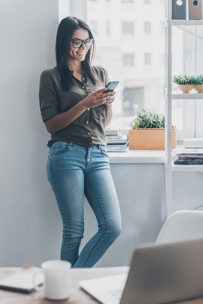 Rimanere in contatto con i colleghi. fiduciosa giovane donna in abbigliamento casual intelligente che tiene in mano uno smartphone e sorride mentre si trova vicino al suo posto di lavoro in ufficio