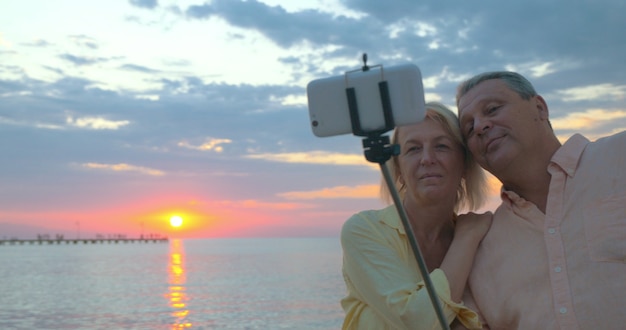Steadicam shot van een senior man en vrouw die buiten mobiele selfie nemen met behulp van selfie stick. ze maken foto's op de achtergrond van zee en zonsondergang en kijken vervolgens naar foto's