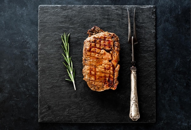 Foto bistecca ribeye al rosmarino su una tavola di ardesia nera e una forchetta di metallo per la carne