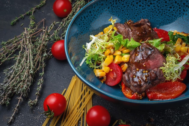 Photo steak salad with vegetables, corn, roasted peppers, cherry tomatoes and soy sauce on a dark stone table