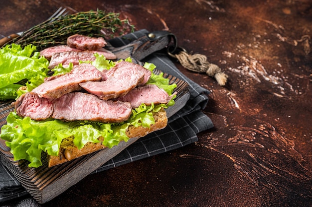 Steak sandwich with sliced roast beef, salad and vegetables. Dark background. Top view. Copy space.