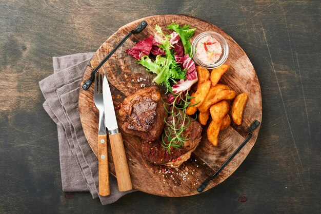 Steaks Sliced grilled meat steak New York Ribeye or Chuck roll with with garnished with salad and french fries on black marble board on old wooden background Top view Mock up