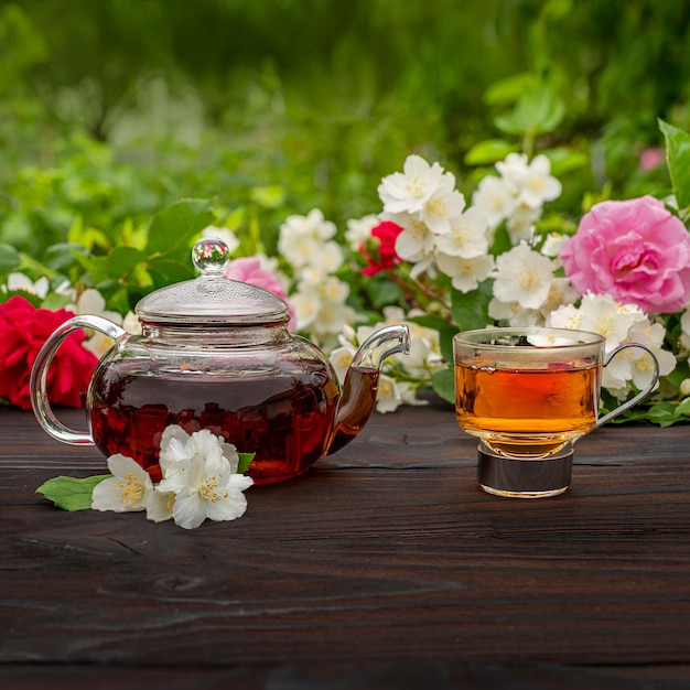 Una teiera al vapore e un'elegante tazza di vetro tra cespugli di rose e gelsomini in fiore