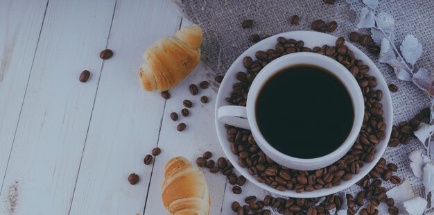 A steaming cup of coffee with coffee beans and a croissant on vintage wooden table