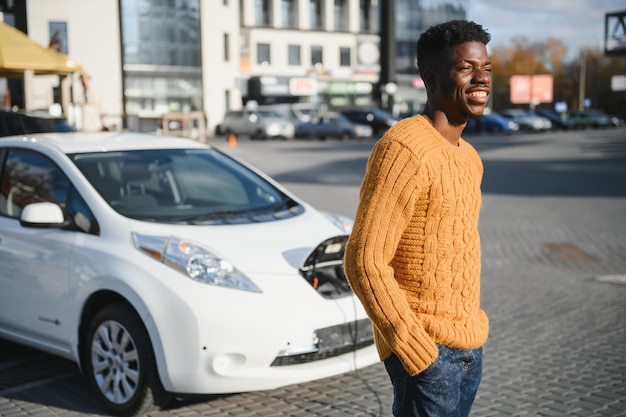 Stedelijk, elektrisch voertuig, eco-concept. Jonge man met zwarte huid, wachtend op het opladen van zijn elektrische auto