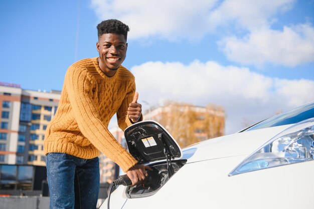 Stedelijk, elektrisch voertuig, eco-concept. Jonge man met zwarte huid, wachtend op het opladen van zijn elektrische auto