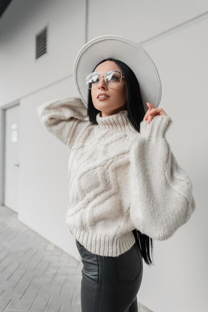 Stedelijk vrouwelijk portret van mooie moderne blanke vrouw met gebreide vintage trui en witte elegante hoed loopt op straat