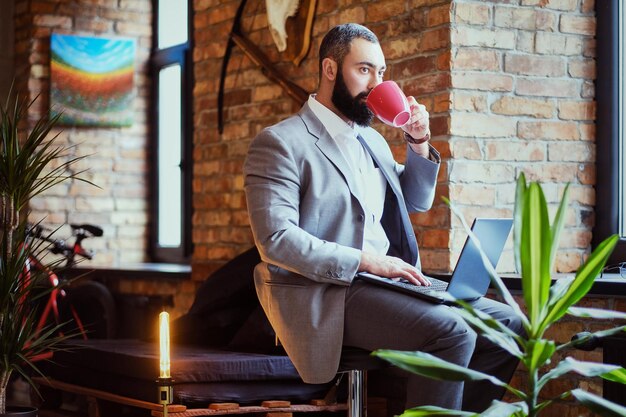 Stedelijke bebaarde man drinkt koffie en gebruikt een laptop in een kamer met loft-interieur.