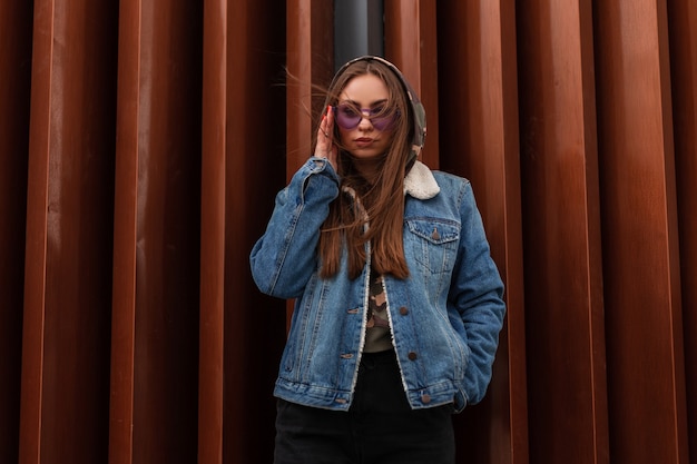 Stedelijke Europese jonge hipster vrouw in jeugd blauw spijkerjasje in paarse mode bril staat in de buurt van rode moderne metalen gestreepte muur in de stad. Aantrekkelijk meisjesmodel in modieuze slijtage buitenshuis. Stijl