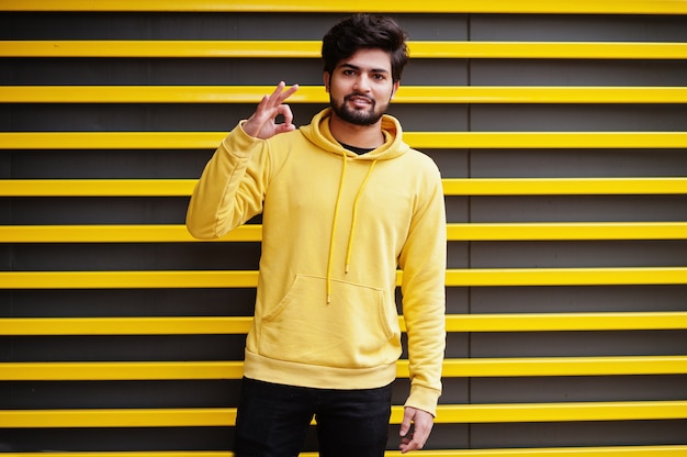 Stedelijke jonge hipster Indiase man in een trendy geel sweatshirt. De koele Zuidaziatische kerel draagt hoodie tegen gestreepte achtergrond die ok toont.