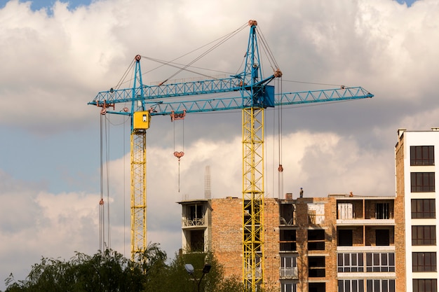 Stedelijke mening van silhouetten van twee hoge industriële torenkranen boven groene boomtoppen die bij bouw van nieuw baksteengebouw werken met arbeiders in bouwvakkers op het tegen heldere blauwe hemelachtergrond.