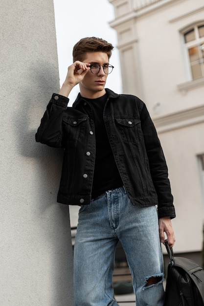 Stedelijke stijlvolle jongeman in denim zwarte jas in spijkerbroek met leren rugzak staat in de buurt van vintage muur en rechtzetten bril. europese knappe man mannequin in modieuze vrijetijdskleding.