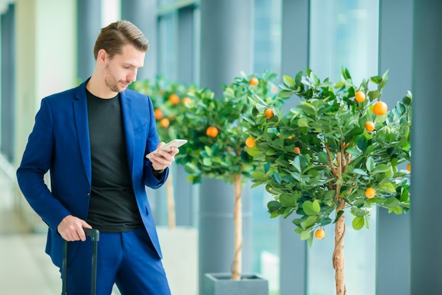 Stedelijke zakenman die op slimme telefoon binnen in luchthaven spreekt. Toevallige jonge jongen die jasje draagt. Blanke man met mobiel op de luchthaven tijdens het wachten op instappen