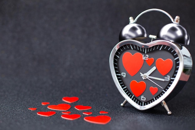 Стальной будильник в виде сердца с красными сердечками внутри и на столе на черном блестящем блестящем фоне С Днем святого Валентина