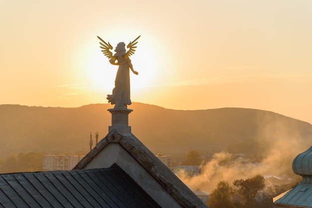 Steenbeeld van een engel met gouden vleugels die bij zonsondergang op het dak van een barokke tempel staat