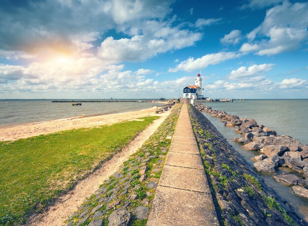 Stenen pad dat leidt naar de vuurtoren aan de zeekust op de achtergrond van blauwe lucht met wolken bij zonsondergang in het voorjaar in Nederland. Landschap met trail, stenen, gras, strand, oceaan en baken. Natuur