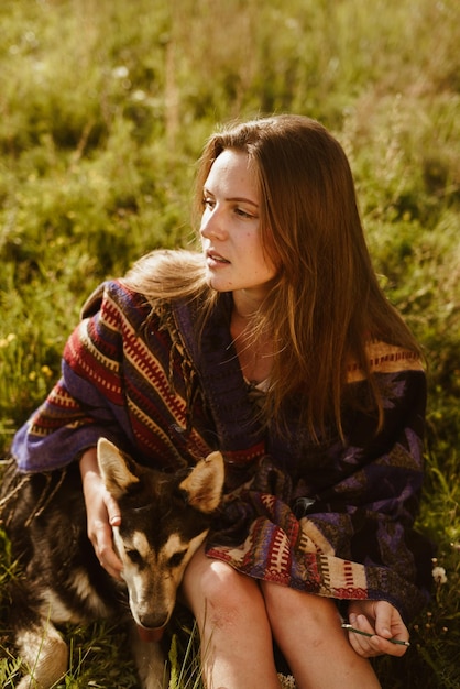 On the steppe grass sits a longhaired blonde beauty with no makeup and hugging the dog looking into the distance