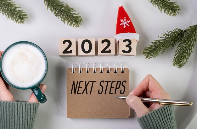 一杯のコーヒーが入ったメモ帳の「次のステップ」の碑文と木製立方体の「2023」の碑文 2023 年の新しい目標のコンセプト