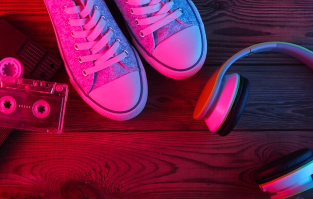 Stereo headphones, audio and video cassette, sneakers on wooden surface. Neon red and blue light