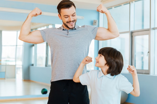 Sterk en gezond. Gelukkige vader en zoon laten hun biceps zien en glimlachen terwijl ze allebei in de gezondheidsclub staan