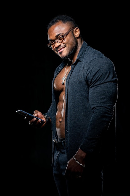 Sterke atleet in donkere spijkerbroek Losgeknoopt zwart shirt en perfecte buikspieren Portret van een Afro-Amerikaanse bodybuilder met telefoon in handen