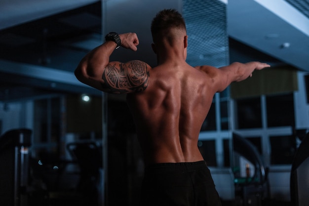 Sterke atletische man bodybuilder met een naakte torso en spieren staat met zijn rug naar de camera in de sportschool in het donker.