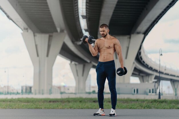 Sterke bodybuilder heft halters op heeft een buitentraining en toont kracht onder het brugfitness- en spiertrainingsconcept