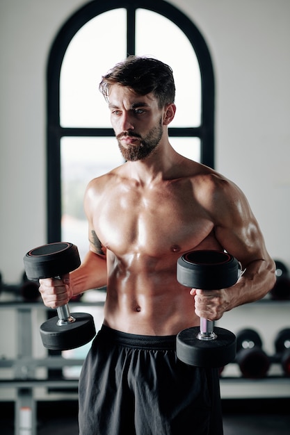 Sterke ernstige shirtless jongeman oefening met zware halters in sportschool