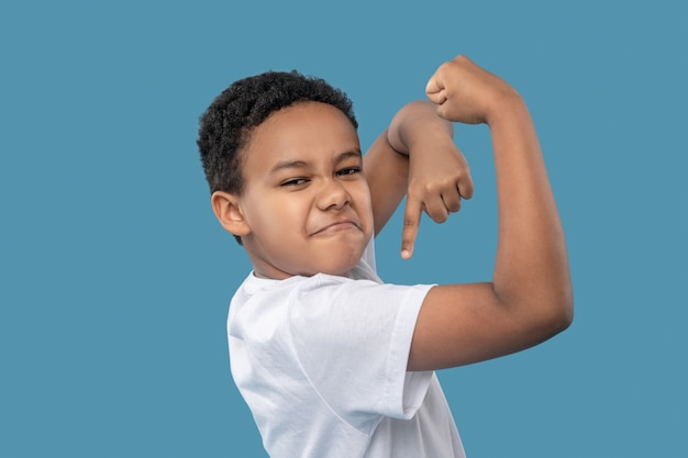 Sterke handen. Grappige schattige jongen met een donkere huid die pronkt met zijn spieren met een vinger die naar de hand wijst en in de studiofoto staat