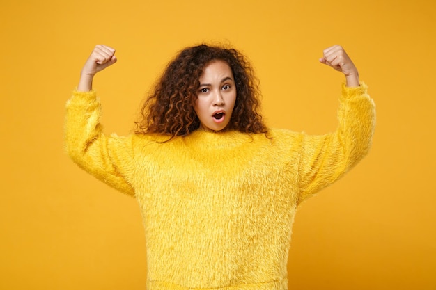 Sterke jonge Afro-Amerikaanse meisje in bont trui poseren geïsoleerd op geel oranje achtergrond, studio portret. Mensen oprechte emoties levensstijl concept. Bespotten kopie ruimte. Het tonen van biceps, spieren.