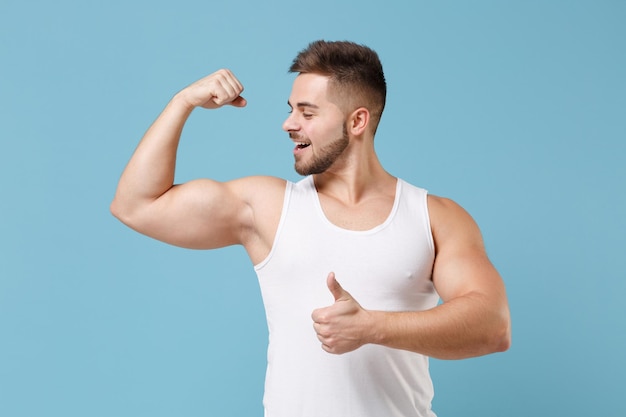 Sterke jonge bebaarde man 20s in wit hemd poseren geïsoleerd op pastel blauwe achtergrond studio portret. Sport fitness gezonde levensstijl concept. Bespotten kopie ruimte. Het tonen van biceps spieren, duim omhoog.