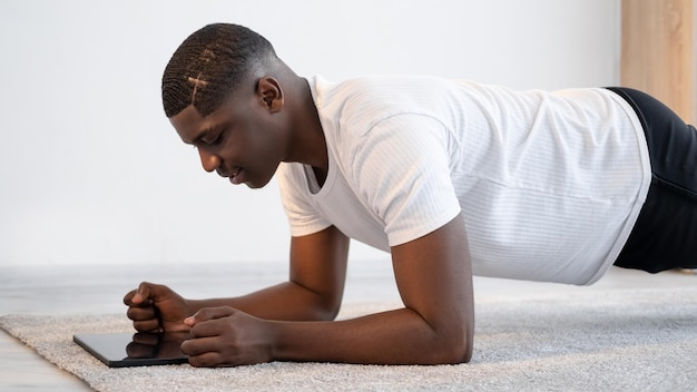 Sterke lichaamstraining Zwarte man Online fitness Thuistraining Gelukkige Afrikaanse man die plankoefening doet terwijl hij op zoek is naar een licht kamerinterieur van een draagbare computer