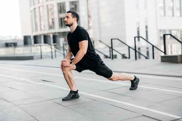 Sterke man die naar voren springt. Zijaanzicht. Sport in grote stad concept. Fit man benen strekken voor het joggen. Runner in zwarte sportkleding opwarmen voor de training. Stedelijk sportconcept.