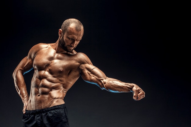 Sterke man met perfecte buikspieren, schouders, biceps, triceps en borst. bodybuilder topless buigen zijn spieren over zwarte muur.