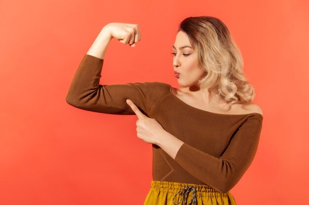 Sterke onafhankelijke vrouw met blond haar die haar armspier laat zien en met de vinger wijst, trots op haar kracht en leiderschapsvaardigheden, emancipatie. Indoor studio-opname geïsoleerd op rode achtergrond