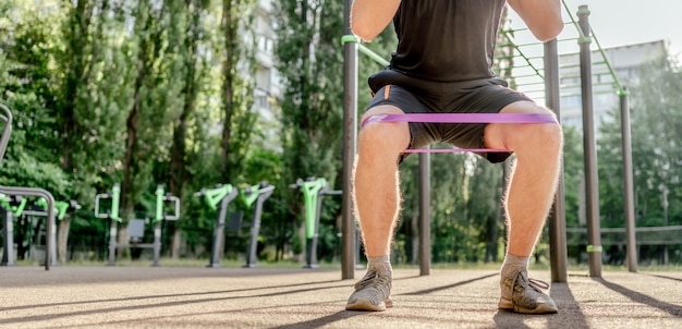 Sterke sportieve man die squats doet met elastische rubberen band in het stadion buitenshuis. Volwassen mannelijke persoon tijdens training voor benen met extra sportuitrusting. Sportman die buiten traint