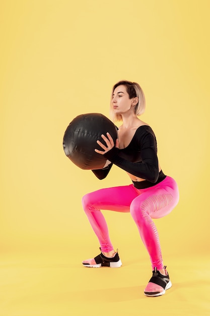 Sterke vrouwentraining met med bal. Foto van sportieve Latijnse vrouw in modieuze sportkleding op gele muur. Kracht en motivatie.