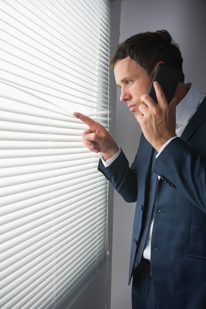 Stern handsome businessman looking through roller blind phoning