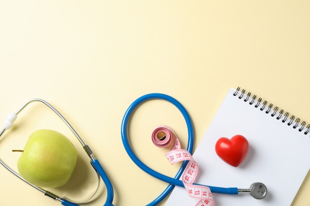 Stethoscope, apple, notebook, heart and measuring tape on yellow background, space for text