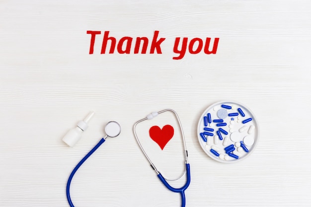 Stethoscope blue colored, pills, red heart and text "thank you" on white wooden background with copy space. Words of gratitude for medical staff.