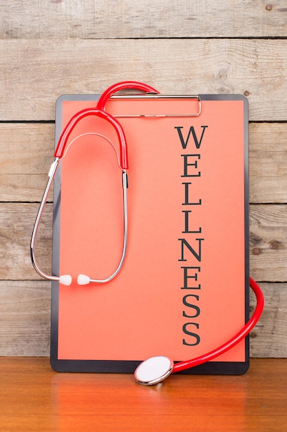 Stethoscope and clipboard with text WELLNESS on wooden desk background