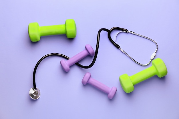 Stethoscope and dumbbells on color background