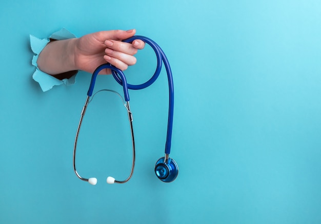 Stethoscope in a female hand on a blue background with a hole, medical concept