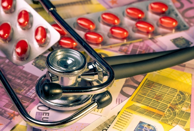 A stethoscope is on top of a pile of money and a pair of pills.