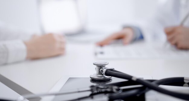 사진 청진기는 의사와 배경에서 서로 반대편에 앉아 있는 환자 앞에 태블릿 컴퓨터에 누워 있습니다. 의학, 의료 개념입니다.